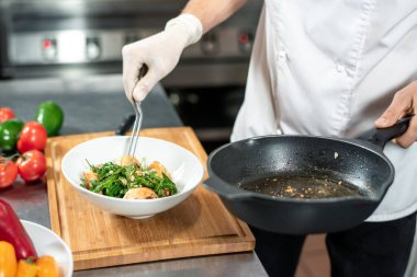 Çatal ve tavayla genç aşçının eldivenli eli büyük masada duran beyaz seramik kaseye pişmiş yemek koyuyor.