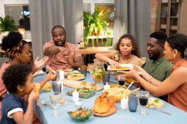 Genç Afrikalı adam fırında pişmiş balkabağı dilimlerini işaret ederken melez bir kadın, bayram masasının yanındaki küçük şirin kıza ızgara mısır uzatıyor.