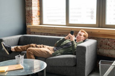 Küçük masa ve büyük ofis penceresi arasındaki yumuşak rahat koltukta dinlenirken akıllı telefon kullanan genç ve dinç bir erkek uzman.