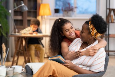 Mutlu Afrikalı kız, dişlek gülüşüyle annesini kitapla kucaklıyor ve arka planda ödevlerini yapan bir çocuğun arka planında kameranın önünde ona bakıyor.