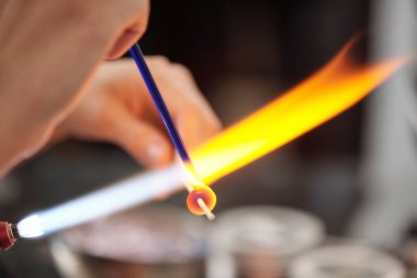 Çağdaş yaratıcı kadın zanaatkar yanan cam işçiliğini ateşe yakın tutarken lambaları yakarken ocaktan çıkıyor.