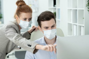 Takım elbiseli ve koruyucu maskeli genç bayan yönetici sunum sırasında erkek meslektaşlarına verileri açıklarken bilgisayar ekranını işaret ediyor.