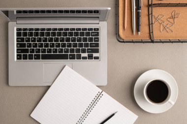 Kahve fincanının en üst görüntüsü, kalem ve boş sayfalarla dolu bir defter, iş yerindeki diğer ofis malzemeleriyle dolu dizüstü bilgisayar ve sepet.