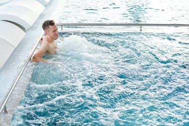 Genç aktif bir adam, arkasında beyaz şezlonglarla yüzme havuzunun kenarında dururken kaynayan suyun tadını çıkarıyor.