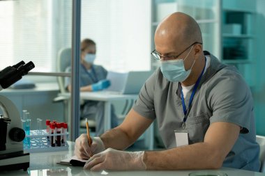 Koruyucu maskeli ve eldivenli çağdaş kel erkek bilim adamı laboratuvar araştırmaları sırasında yeni aşılar hakkında bilimsel notlar alıyor.