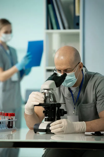 마스크와 장갑을 연구하는 대머리 과학자 현미경으로 샘플을 연구하는 조수에 — 스톡 사진