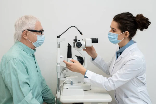 年轻女性眼科医生在临床使用医疗设备检查戴防护面具老年男性患者视力时的侧视 — 图库照片