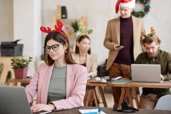 年轻的女办公室经理戴着眼镜 穿着时髦的便服 头戴圣诞帽 站在笔记本电脑前与同事们交流 — 图库照片