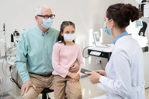 在诊所就诊期间 老先生和他的孙女戴着防护口罩 坐在年轻女性眼科医生的面前 — 图库照片