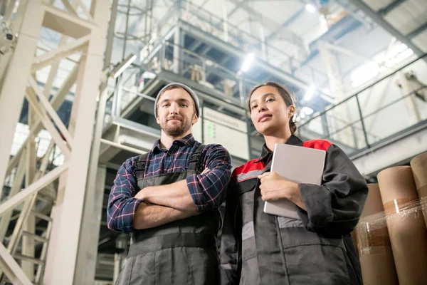 大工厂的年轻成功的男女工程师站在镜头前 用巨大的技术结构看着你 — 图库照片