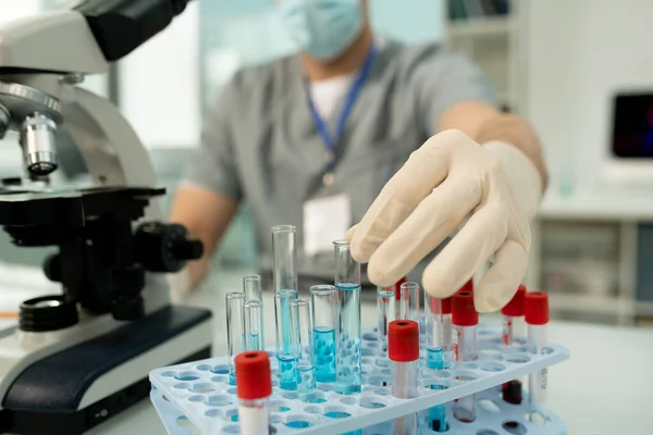 新しいワクチンのテスト中に青色液体化学物質のサンプルとフラスコを取って医療研究所の現代の労働者の手袋の手 — ストック写真