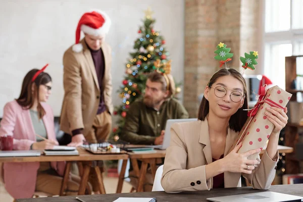 幸せな若いですアジアのビジネスマン女性でXmasヘッドバンド座っていますテーブルと持っています大パックギフトボックスによって彼女の耳を閉じながら — ストック写真