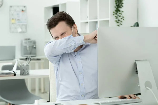 Young male office manager in protective mask putting his face on arm while sneezing by workplace in front of computer monitor during work