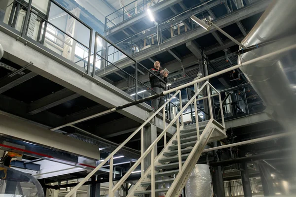 階段の前に現代的な大規模なポリマー加工工場内の技術データを扱うデジタルタブレットと大胆な成熟したフォアマン — ストック写真