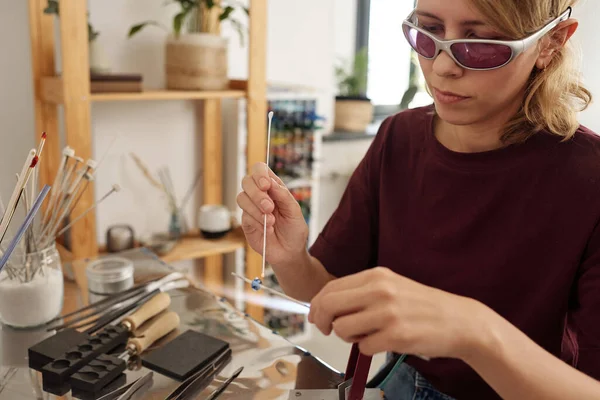 作業場に座っている保護眼鏡の若い女性職人とテーブルの上にそれを保持しながら スティック上のガラスワークを飾る — ストック写真