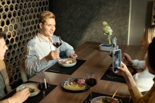 友人の間でモダンなレストランでテーブルに座っている間に夕食で赤ワイン作りトーストのガラスと幸せな若いエレガントな男性 — ストック写真