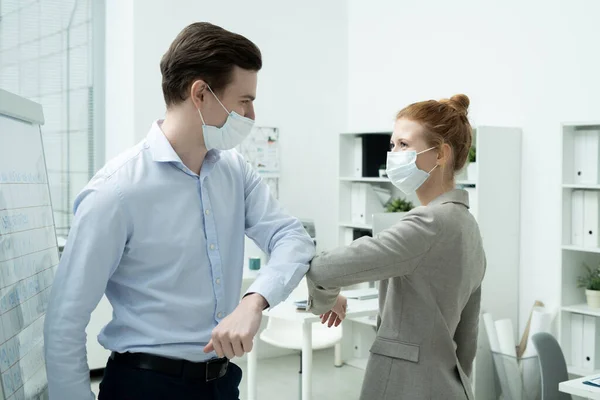 Two young happy office managers in formalwear and protective masks giving each other elbow bump instead of handshake or hug