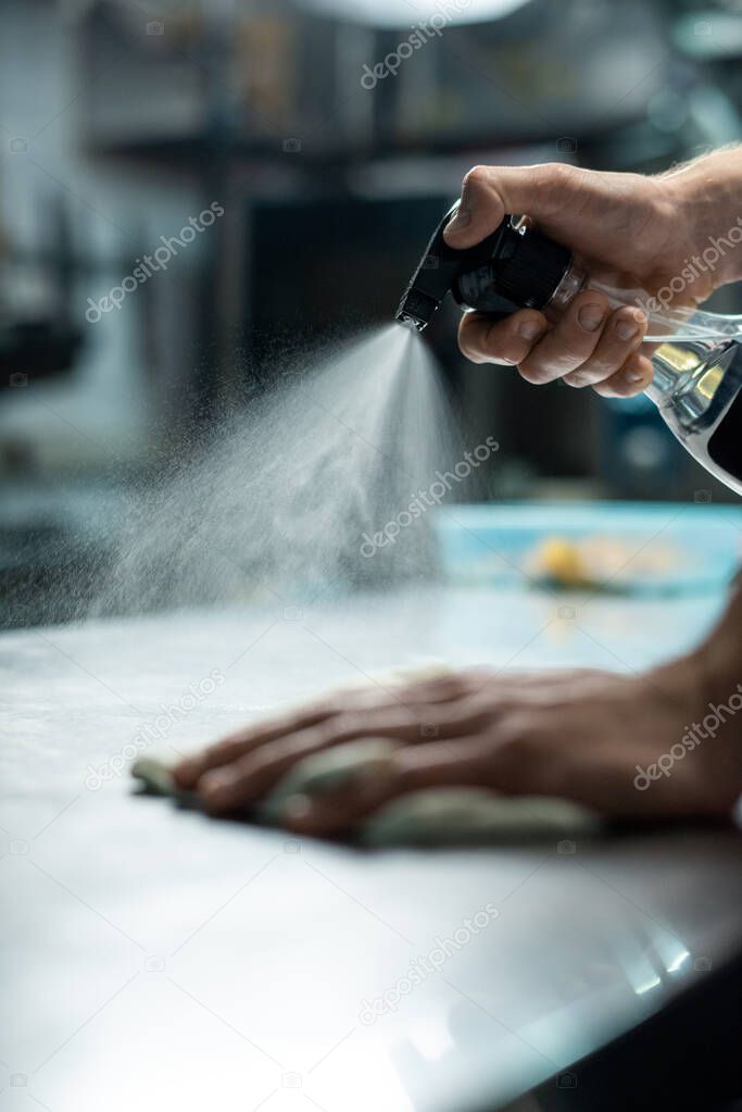 Hands of young contemporary male worker of restaurant kitchen spraying sanitizer on table while cleaning it before preparation of food