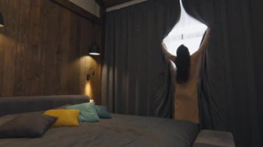 Uzun elbiseli zarif esmer kadının, lüks otel odasının karanlık perdelerini açarken, manzaranın keyfini çıkardığı yavaş çekimde.