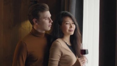 Havalı bir otel odasının penceresinde kameraya gülümseyen, elinde kırmızı şarap kadehi olan romantik, çok ırklı bir çiftin göğüs hizasında ağır çekimde portresi.