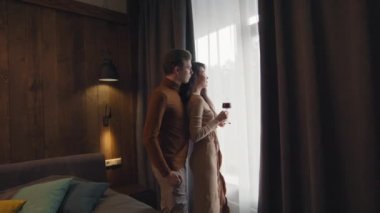 Karanlık bir otel odasının penceresinde elinde bir bardak kırmızı şarapla bekleyen, sevgi dolu, çok ırklı bir çiftin orta boy portresi.