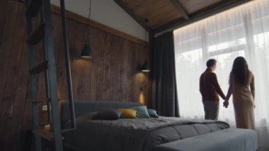 Arka planda, şık bir otel odasında pencerede durup el ele tutuşan ve pencereye bakan romantik bir çiftin görüntüleri var.