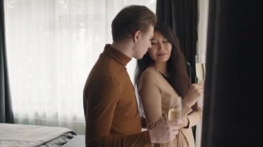 Otel odasında şampanya bardakları olan, eşine arkadan sarılan romantik, çok ırklı bir çiftin orta boy yakın çekimi.