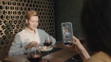 Omzunun üstünden erkek arkadaşının fotoğrafını çeken bir kadın restorandaki masada bir bardak kırmızı şarapla akıllı telefondan fotoğraf çekiyor.
