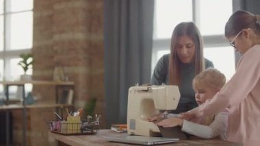 5 yaşındaki küçük bir kızın dikiş makinesiyle dikiş dikmeyi öğrendiği orta boy bir fotoğraf. Oturma odasında oturuyor ve annem ve ablam onun yanında durup...