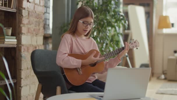 中出しの12歳の女の子の椅子に座って自宅でオンライン音楽レッスンを持っていますラップトップで演奏ウクレレを練習 — ストック動画