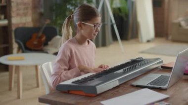 Küçük mutlu kızın evde dizüstü bilgisayarla müzik dersi alıp klavye çalmayı öğrendiği orta boy bir fotoğraf.