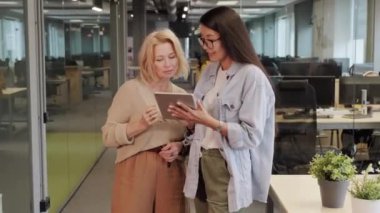 Orta yaşlı beyaz kadın şirket CEO 'sunun modern açık uzay ofisinin ortasında Asyalı bir ofis çalışanı ile birlikte tablet üzerine yeni projenin sunumunu kontrol ederken orta boy fotoğrafı.