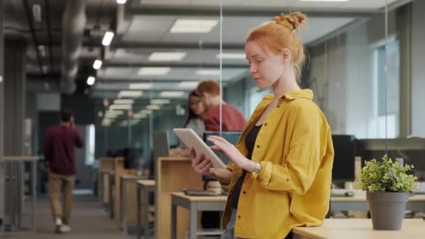 中慢镜头拍摄的红头发年轻的办公室女职员穿着鲜艳的黄色衬衫站在宽敞的大办公室的平板电脑上 而同事们则在后台交流 — 图库视频影像