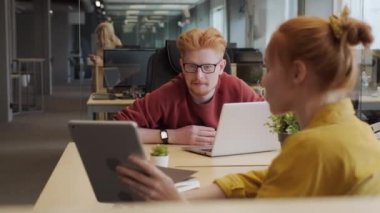 İki gülümseyen kızıl saçlı ofis çalışanının tablet ekrana bakıp yeni projeyi tartıştığı orta boy yakın plan parlak ve ferah ofisteki işyerlerinde otururken.