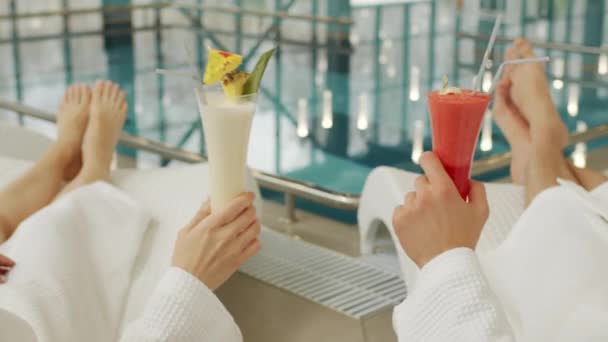 美しいカクテルと屋内プールのクリンクグラスでラウンジャーに横たわっている白いバスローブの認識できないカップルのクローズアップ — ストック動画