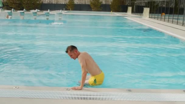 バックビュースローモーションショットの若いですハンサムな男がプールの水から出て来て 楽しい時間を持ってプールサイドに座って — ストック動画