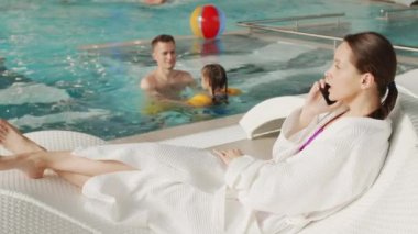 Yavaş çekimde, genç mutlu aile termal havuzda rahatlarken, baba suda kızıyla oynarken anne telefonla konuşurken.