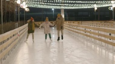 Anne, baba ve küçük kızın soğuk kış akşamlarında boş buz pateni sahasında buz pateni yaparken çekilmiş neşeli bir aile görüntüsü.