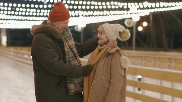 寒い冬の夜に屋外のアイスリンクで話す冬服のロマンチックな高齢者カップルのウエストアップ映像 — ストック動画