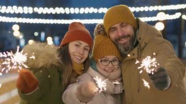 Akşamları buz pateni pistinde kameraya gülümseyen üç kişilik gülümseyen beyaz bir ailenin orta boy portresi.