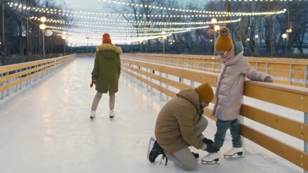 フルショットの男結ぶ靴ひも上の小さな娘フィギュアスケートその後結合お母さんとスケート一緒に手を持っています空オープンエアアイスリンク — ストック動画