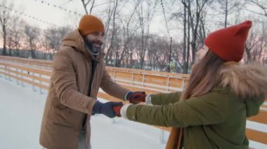 Bir adamın sevgilisine kışın gün batımında buz pateni pistinde buz pateni öğretirken orta ölçekli bir görüntüsü.