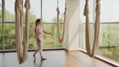 Dar pastel, pembe spor kıyafetli genç bir kadının Aero Yoga Stüdyosu 'na girmesi ve yoga sapanını tutarak egzersiz yapmaya başlaması.