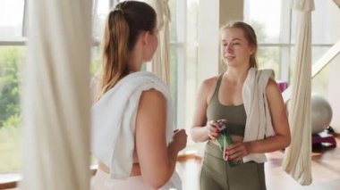 Spor kıyafetlerinde sohbet eden iki kız arkadaşın el kamerasıyla çekilmiş resmi. Panoramik pencereli bir yoga stüdyosunun önünde dikiliyorlar.