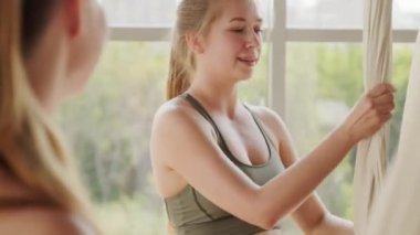 Aero yogası için ipek hamak serpiştiren ve panoramik pencereli, parlak bir odada duran bayan arkadaşıyla sohbet eden beyaz bir kadın.