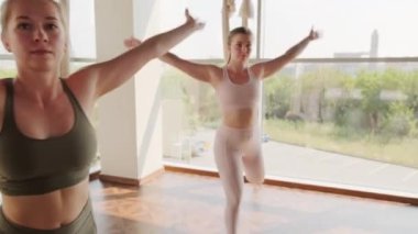 Sıkı spor giyim giymiş, yoga yapan bir grup beyaz kadının bir ayağı üzerinde, bir ayağı hamağın arkasında ve elleri dua pozisyonunda dengede durduğu yavaş çekim görüntüleri.
