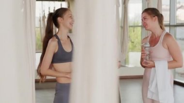 Üç zayıf beyaz kızın spor salonu yoga eğitiminden sonra sohbet ederken çekilmiş orta boy portresi. Elinde form şişeleriyle kameraya bakıp gülümsüyor.