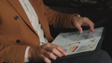 Araba kullanırken arabanın arka koltuğunda oturan deve ceketli tanınmış işadamının orta kesiminde baskılı çizelgeler ve diyagramlar inceleniyor.