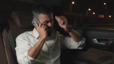 Beyaz tişörtlü, melez bir iş adamının gecenin bir yarısı taksinin arka koltuğunda telefonla konuşurken, bağırırken ve buluşmaya geç kalırken orta boy görüntüsü.