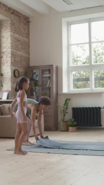 Küçük anne ve kızının spor kıyafetleri içinde yoga minderinde otururken ve oturma odasında birlikte pratik yaparken dikey çekimleri.